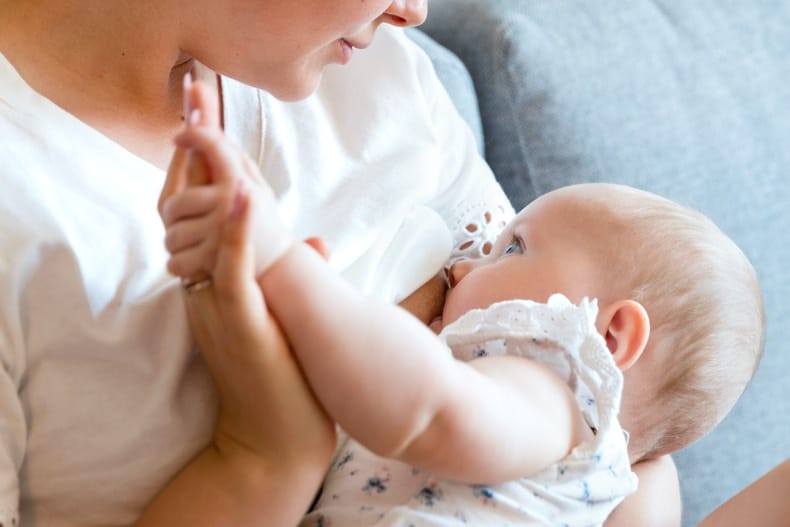 A mother is holding her baby at her breast, and gazing down at baby while they gaze up at Mom and feed. Mom and baby's hands are also tenderly clasped together.