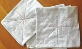 overhead shot of cloth baby wipes on a wooden surface. wipes are white fabric squares with red thread sewn in a starburst pattern.