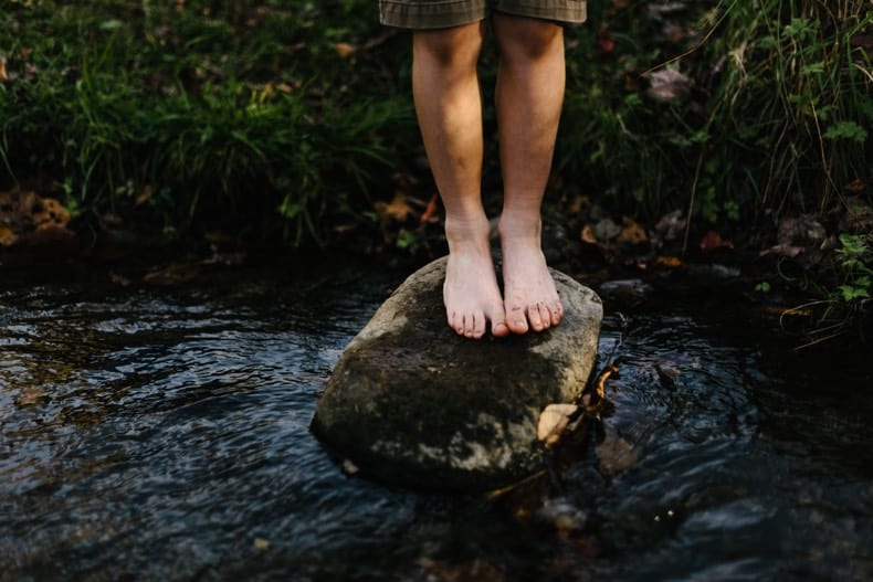 a person from the knees down, standing barefoot on a rock in a stream, enjoying grounding with nature